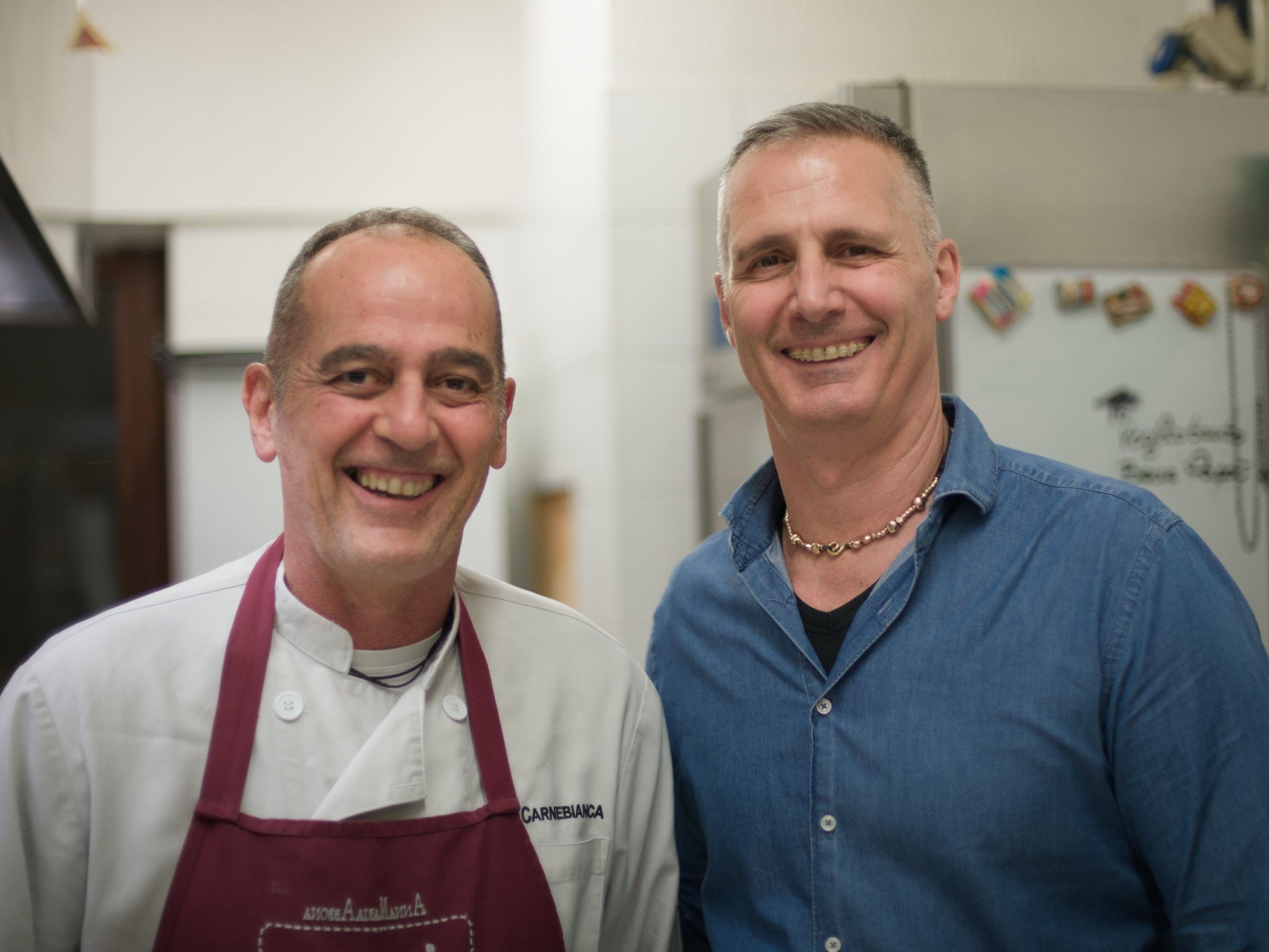 Fabio, con il grembiule rosso da cuoco, e Christian, in camicia di jeans, sorridenti nella cucina dell'osteria