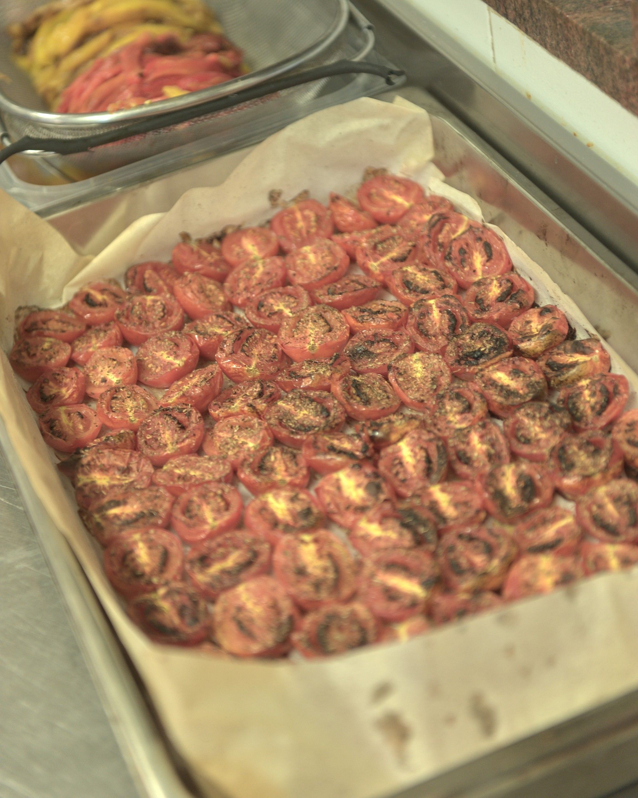 Una teglia di pomodorini tagliati a metà e arrostiti al forno, con i peperoni arrostiti nel colino dietro