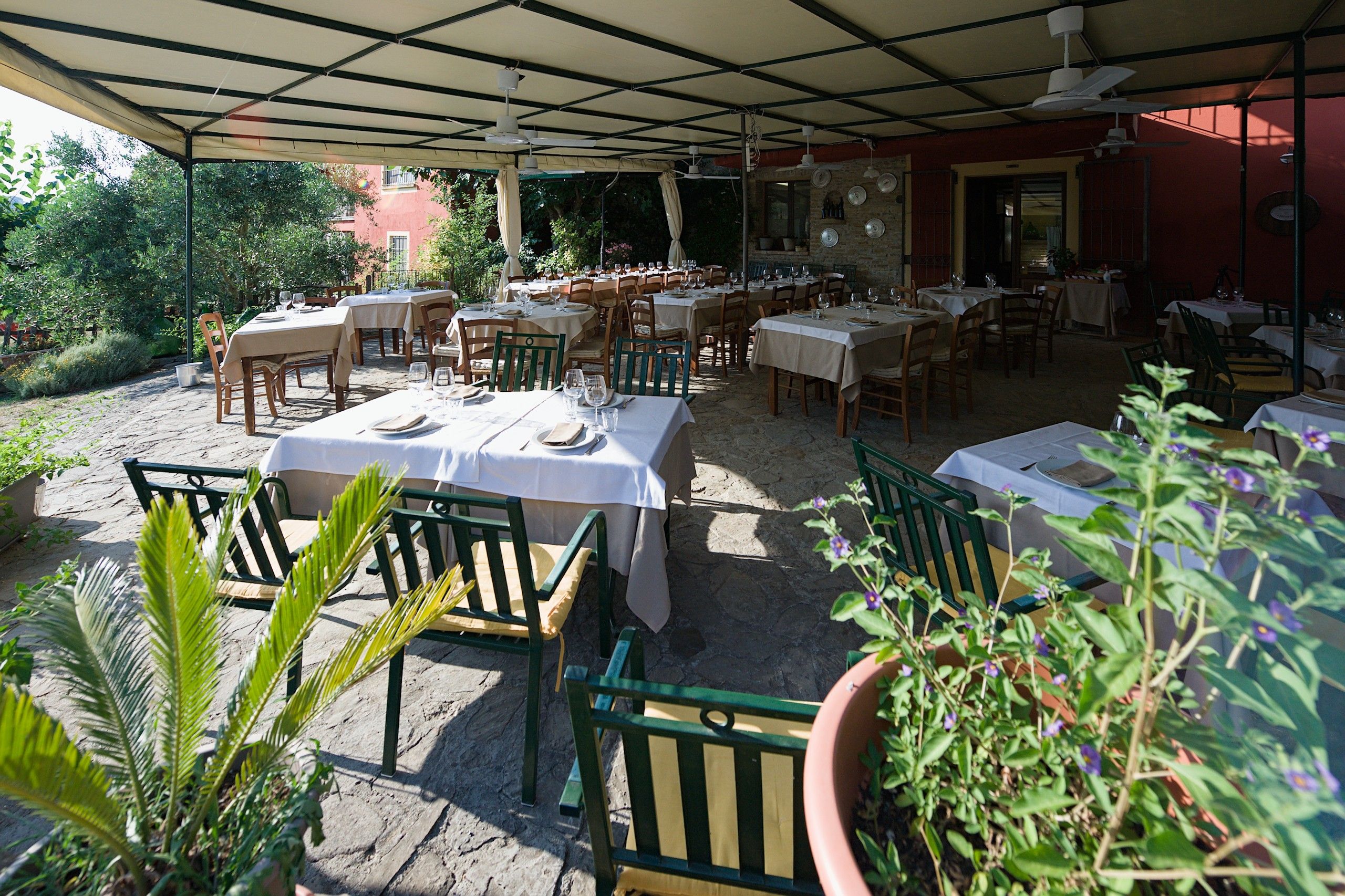 La terrazza coperta con i tavoli apparecchiati, le sedie verdi e le piante in vaso, con il muro in pietra e i piatti appesi sullo sfondo